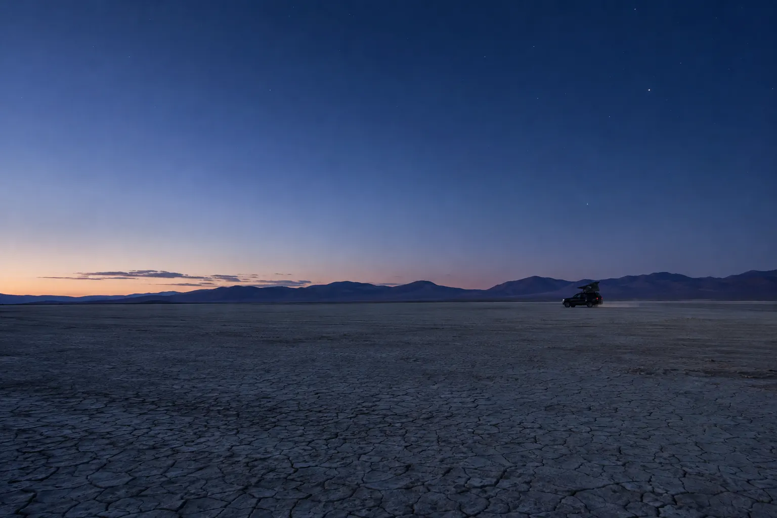 A lone vehicle on a cracked desert playa at dusk with distant mountains